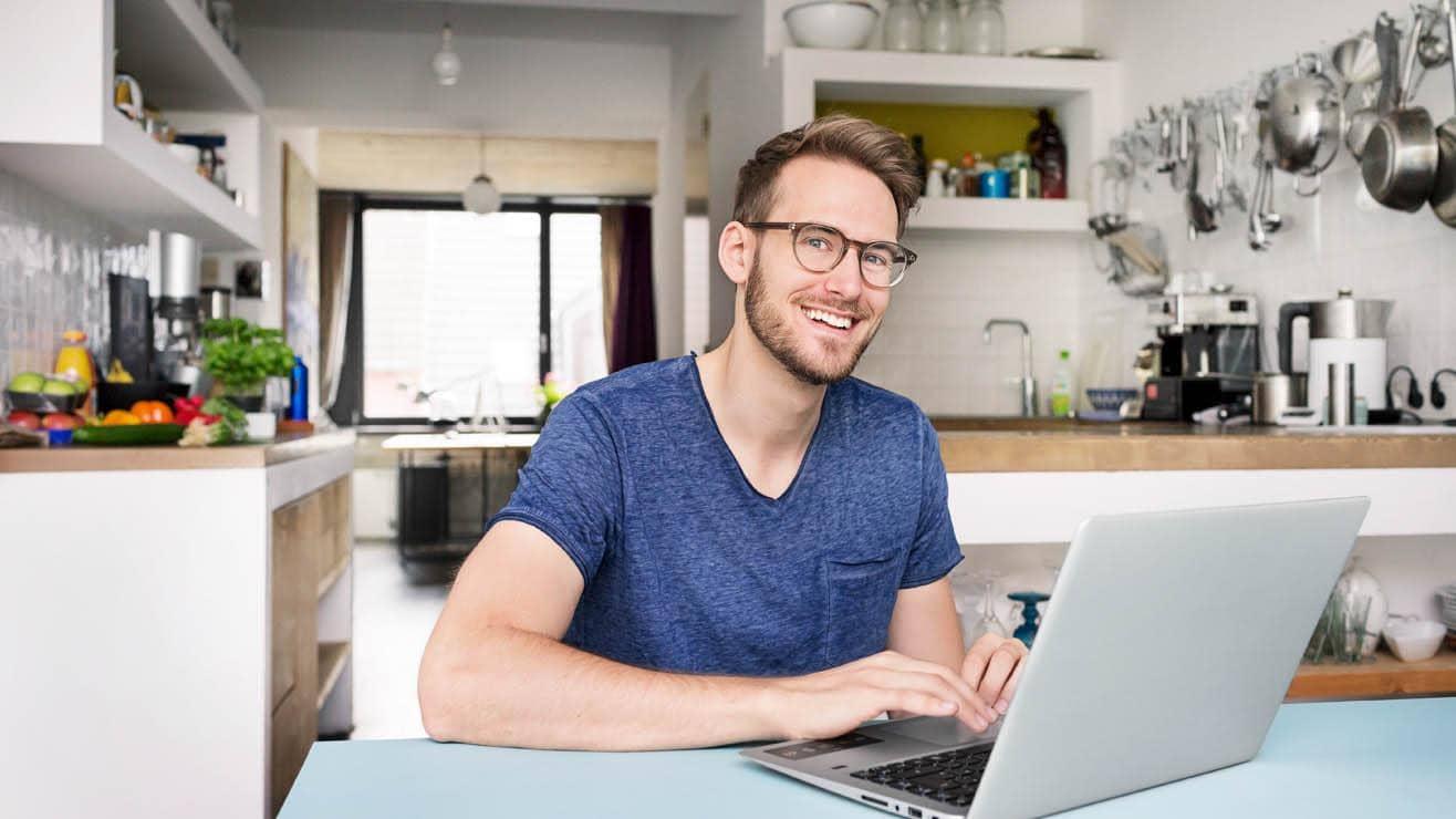  A man sitting behind a laptop in his kitchen smiles at the camera